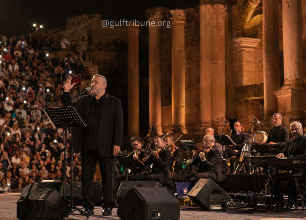 Male singer in black suit performing on stage with a microphone, backed by an orchestra at night near ancient columns; large audience in the stands.