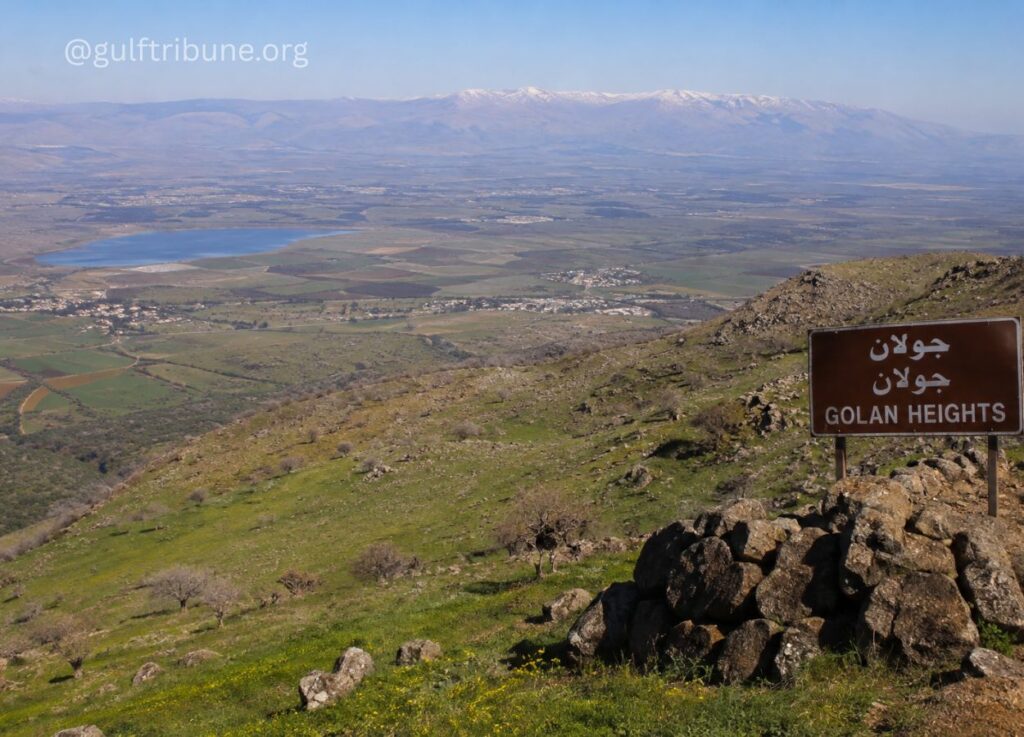 View from a hillside at Golan Heights overlooking fields, a lake, and distant mountains; sign reads 'Golan Heights' in Arabic and English.