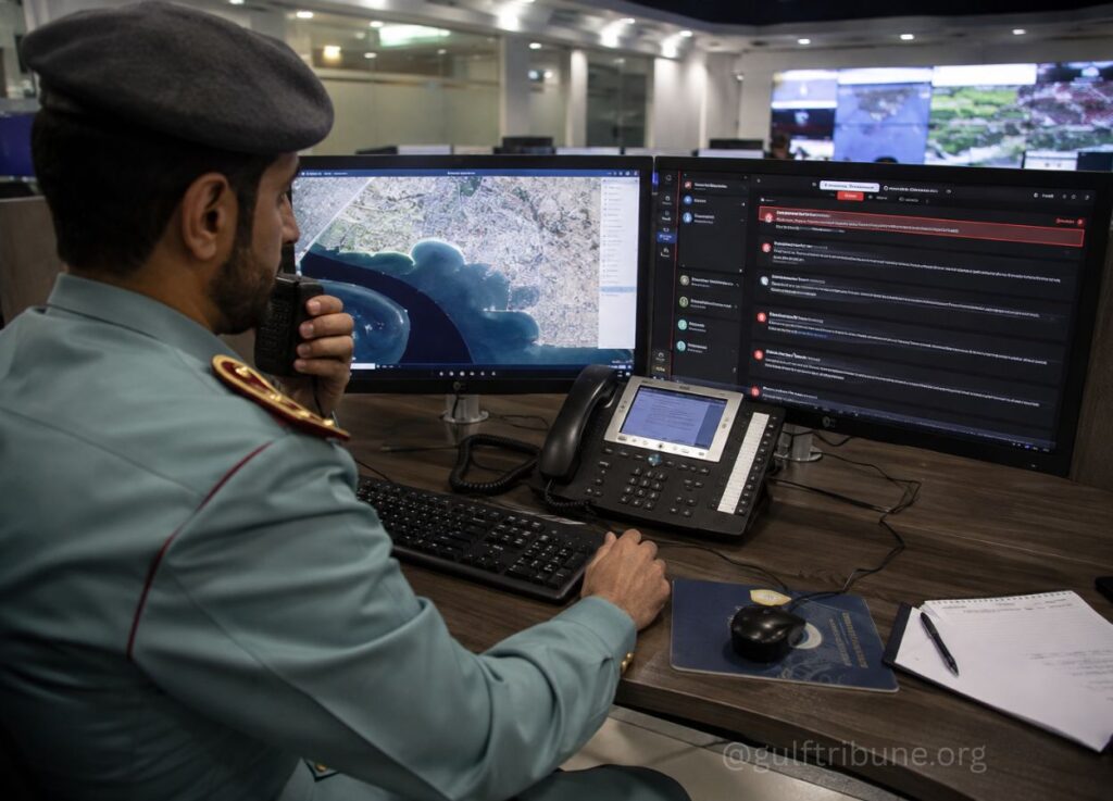 Uniformed officer in a command center speaks into a handheld radio while monitoring a map on dual computer monitors.