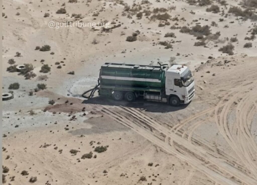 144 - Gulf Tribune White tanker truck with a green cylindrical tank sits on a sandy desert site, with tire tracks and a circular wet patch nearby.