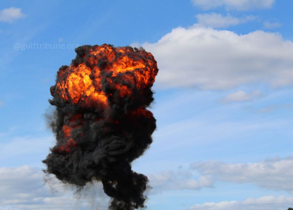 Massive fiery explosion with orange flames and dark smoke rising into a blue sky.