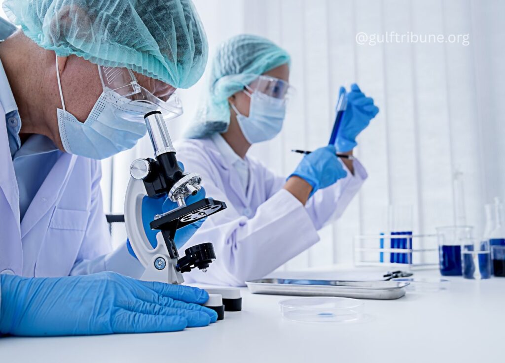 77 - Gulf Tribune Two scientists in protective gear use a microscope and pipette in a modern laboratory site.