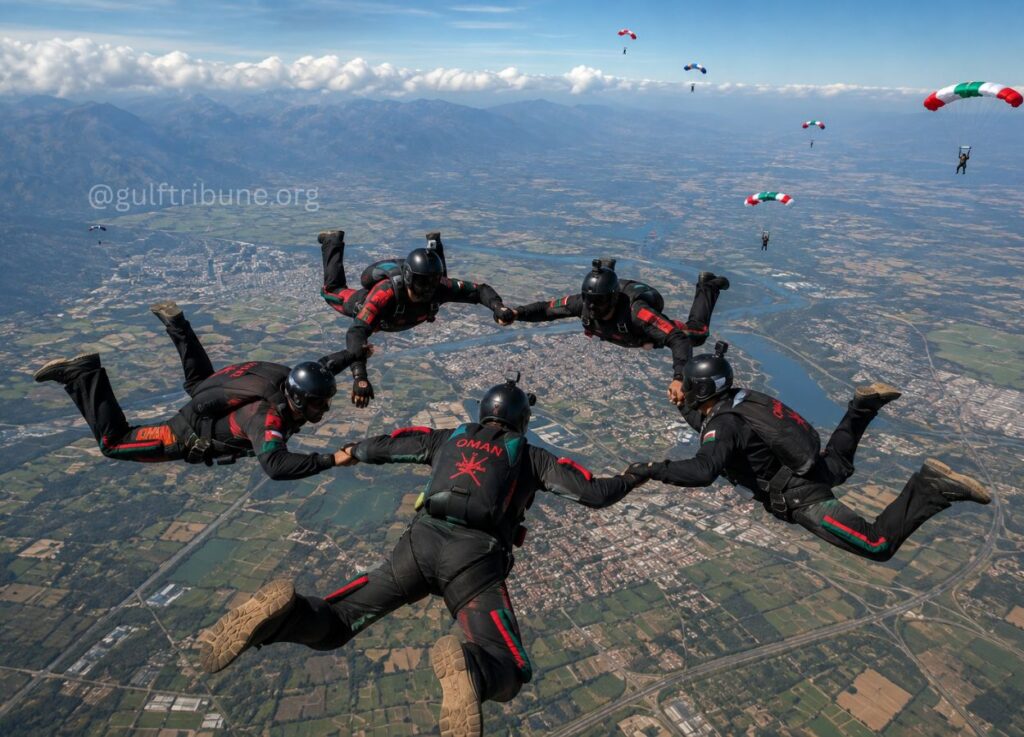 Six skydivers in black and red suits form a circle, holding hands in freefall above a patchwork valley with fields and a river.