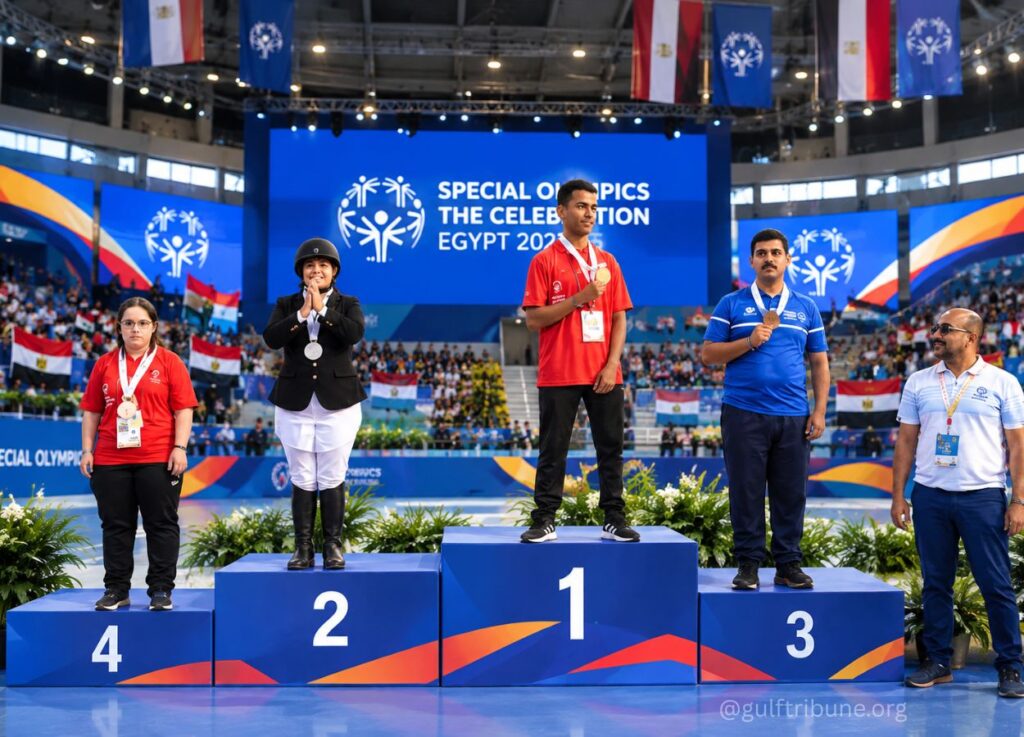 Four athletes stand on a blue podium with numbers 1–4 during a Special Olympics awards ceremony in a packed indoor arena; the gold medalist stands on the center podium, silver on the left, bronze on the right, with a crowd, flags, and a large blue screen that reads